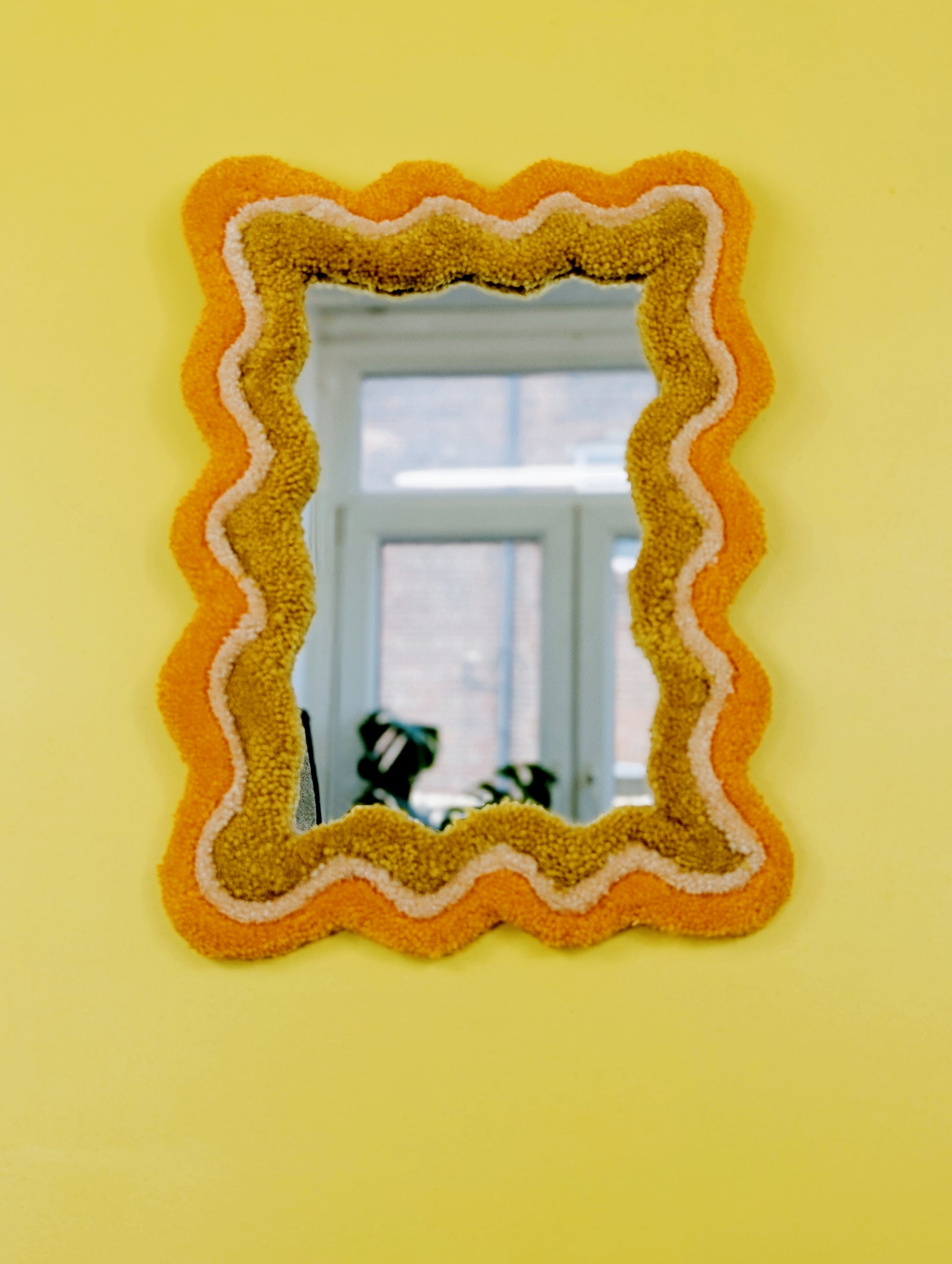 Handmade wavy tufted mirror with colourful textured frame in alternating shades of orange, cream and mustard yellow, modern aesthetic wall décor for home interiors.