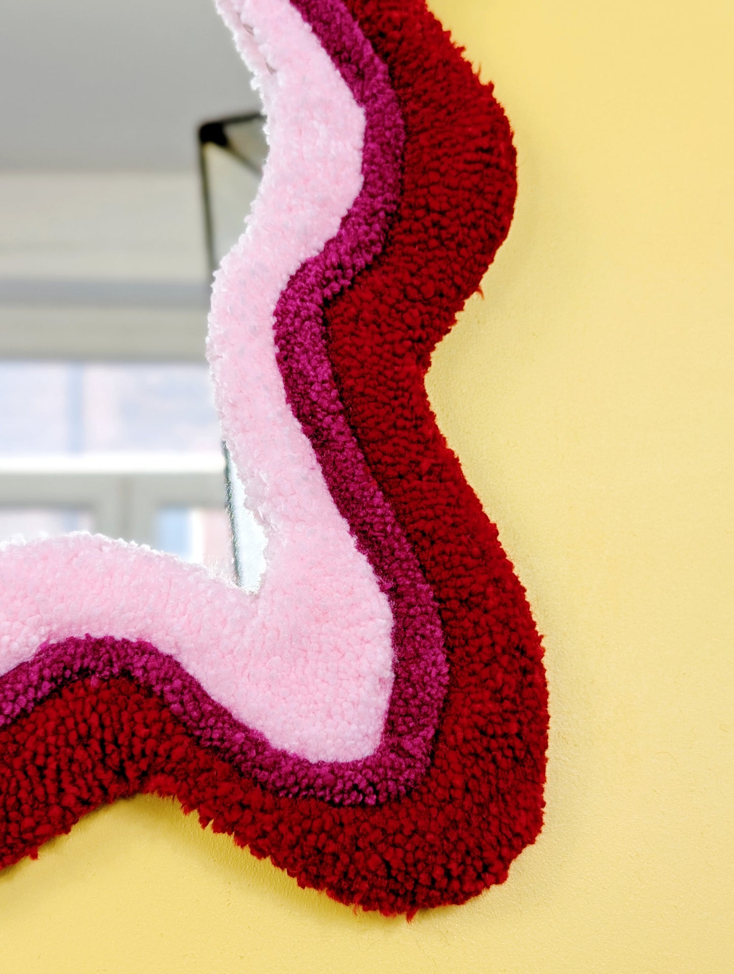 Handmade wavy tufted mirror with colourful textured frame in alternating shades of bright red, dark pink and pale pink, modern aesthetic wall décor for home interiors.
