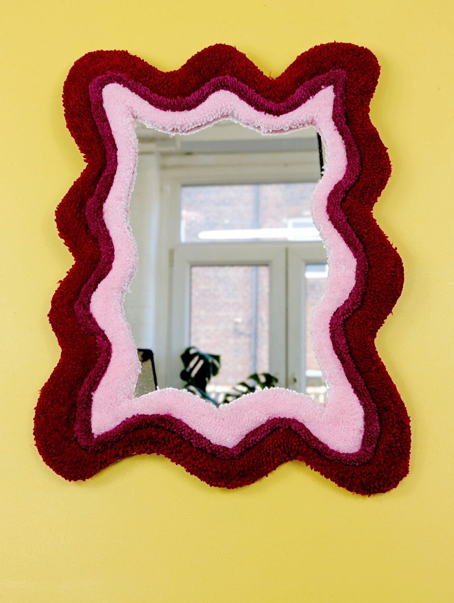 Handmade wavy tufted mirror with colourful textured frame in alternating shades of bright red, dark pink and pale pink, modern aesthetic wall décor for home interiors.