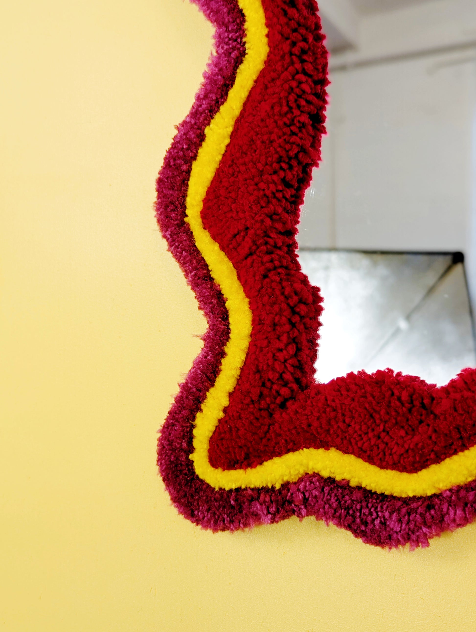 Handmade wavy tufted mirror with colourful textured frame in dark pink, bright yellow and red, modern aesthetic wall décor for home interiors.