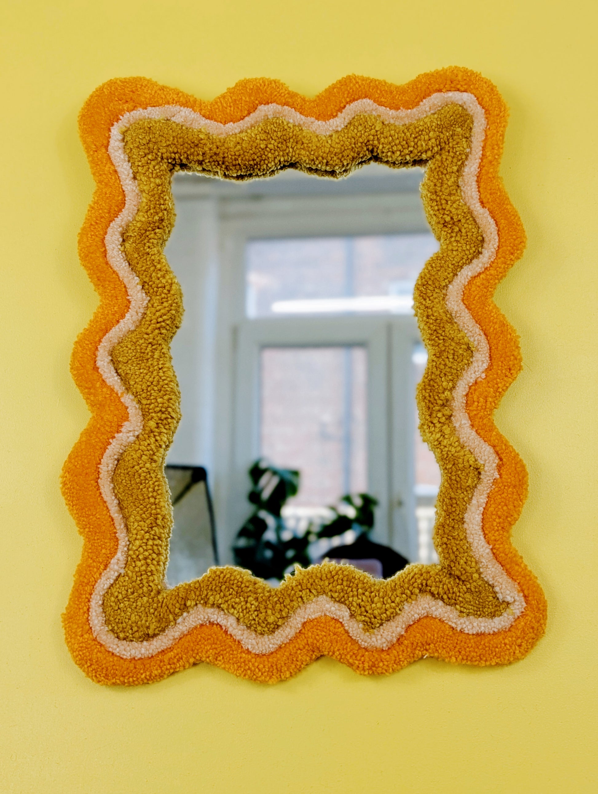 Handmade wavy tufted mirror with colourful textured frame in alternating shades of orange, cream and mustard yellow, modern aesthetic wall décor for home interiors.