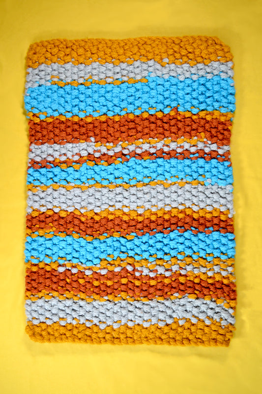 Hand knitted rug on a yellow background. Rug has a striped pattern. Rug is mustard yellow, burnt orange, grey and crisp sky blue stripes, with thick mustard yellow stripes at both ends. Each individual stitch is visibly and the rug has an extra thick pile height.