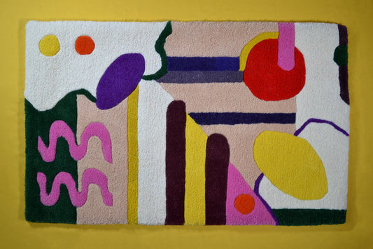 Image shows a cream rug with multicoloured abstract shapes on it. These shapes are red, burgundy, sunflower yellow, hot pink, dark green and orange. the background of the rug has 2 beige block colour sections. The rug is photographed against a yellow background. 
